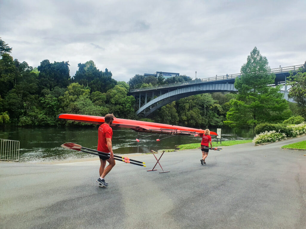 Shane Quintus and another member of the club carry a boat to Waikato River with Victoria Bridge in the background.